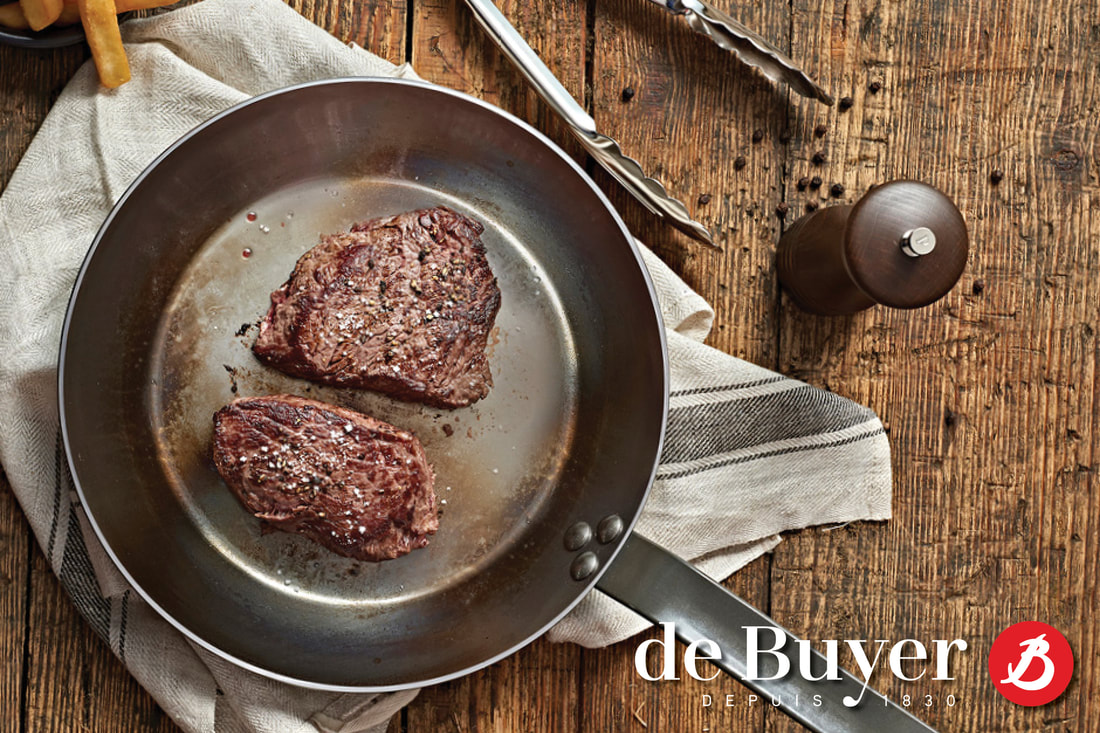 de Buyer fry pan with grilled steak , tongs and a pepper mill on a table. 