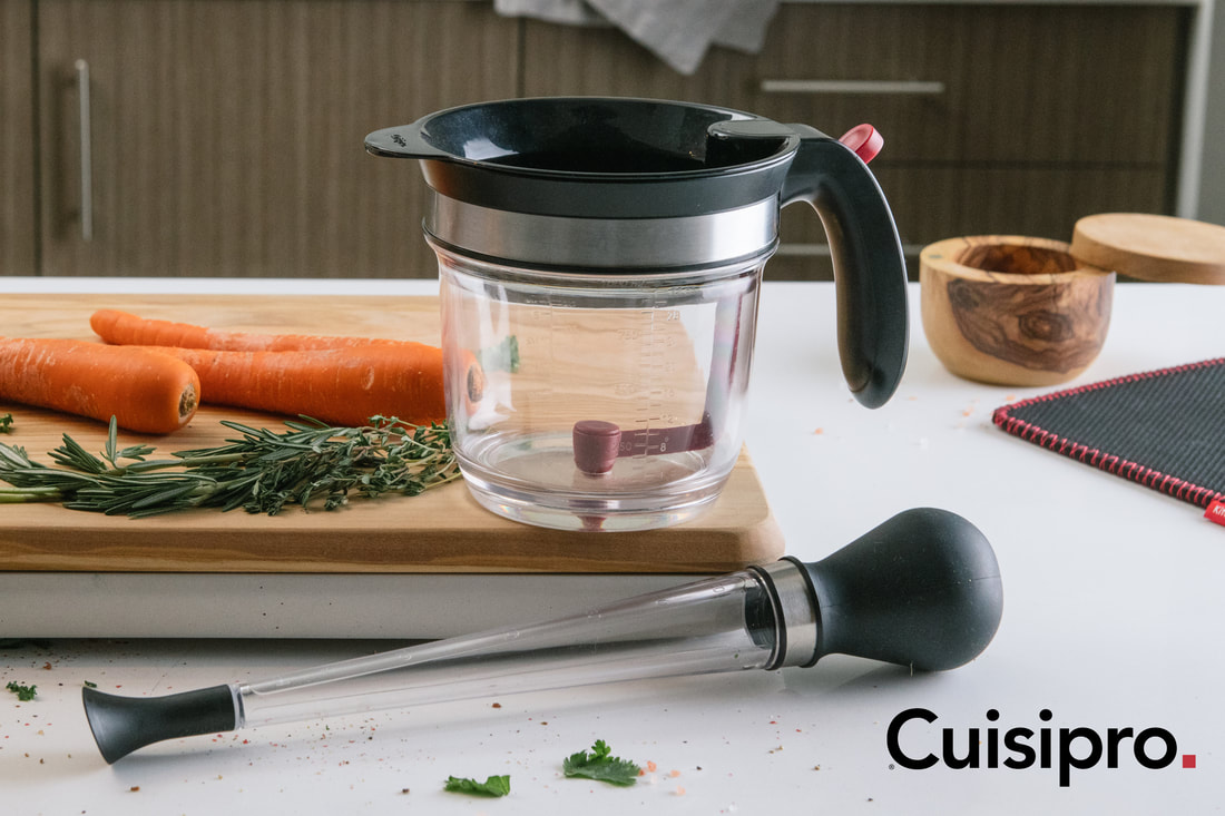 Cuisipro fat separator and baster in kitchen with wood cutting board, carrots and herbs. 